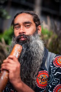 A bearded man playing a didgeridoo, wearing culturally patterned clothing, outdoors in Sydney.